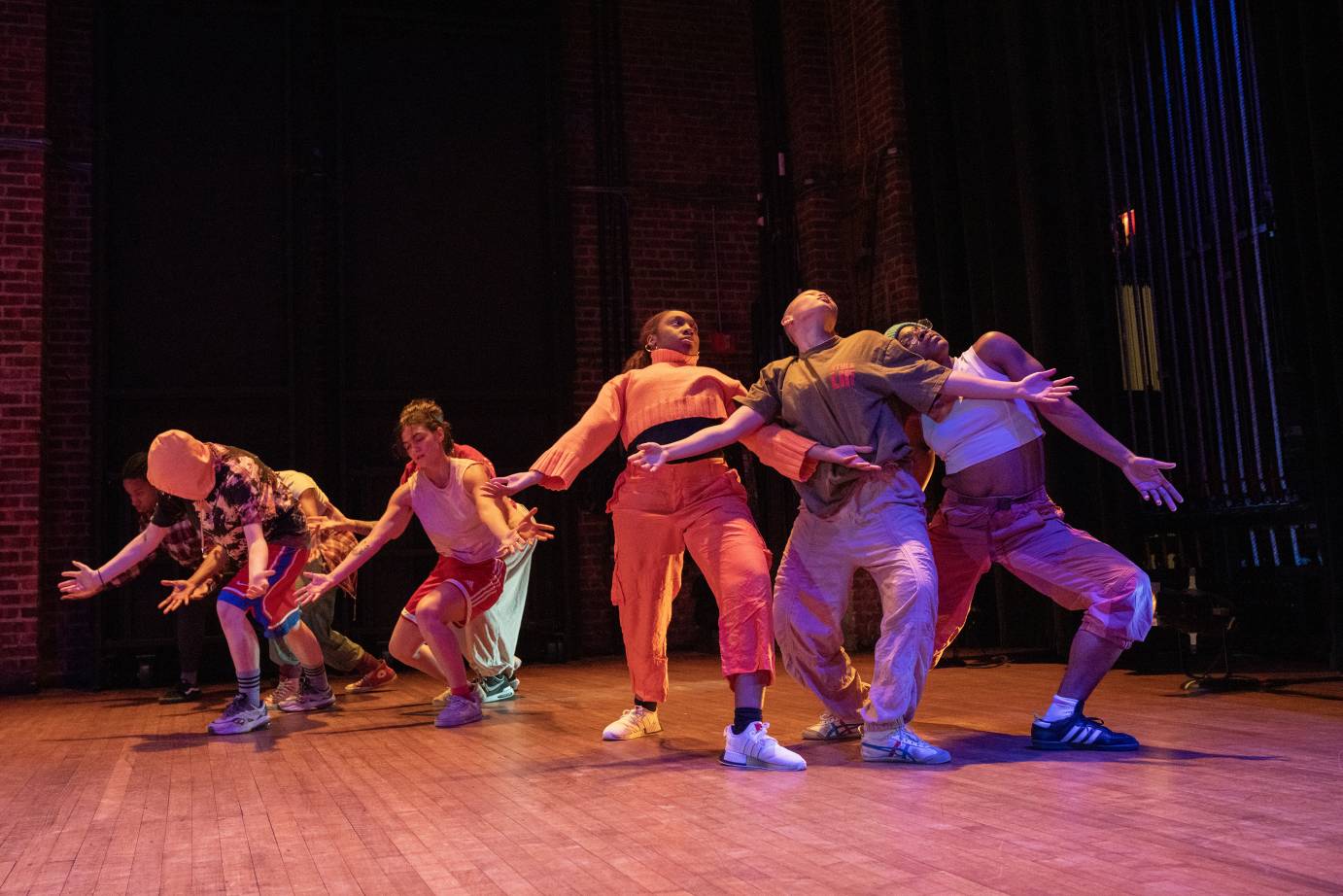 Two groups of young dancers in colorful street gear share the stage. The first group leans forward towards the floor with arms streched in front of them. The second group, a trio, leans into a back-bend as a group, they support one another with their arms and torsos while their legs are bent.